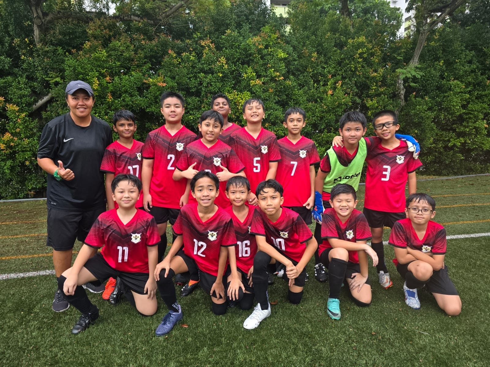 A youth soccer team in red and black jerseys poses with a coach on a grass field in front of green trees.