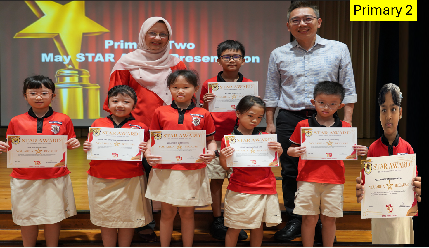 Seven young students in red uniforms hold Star Award certificates, posed with two adults at a Primary 2 award ceremony.