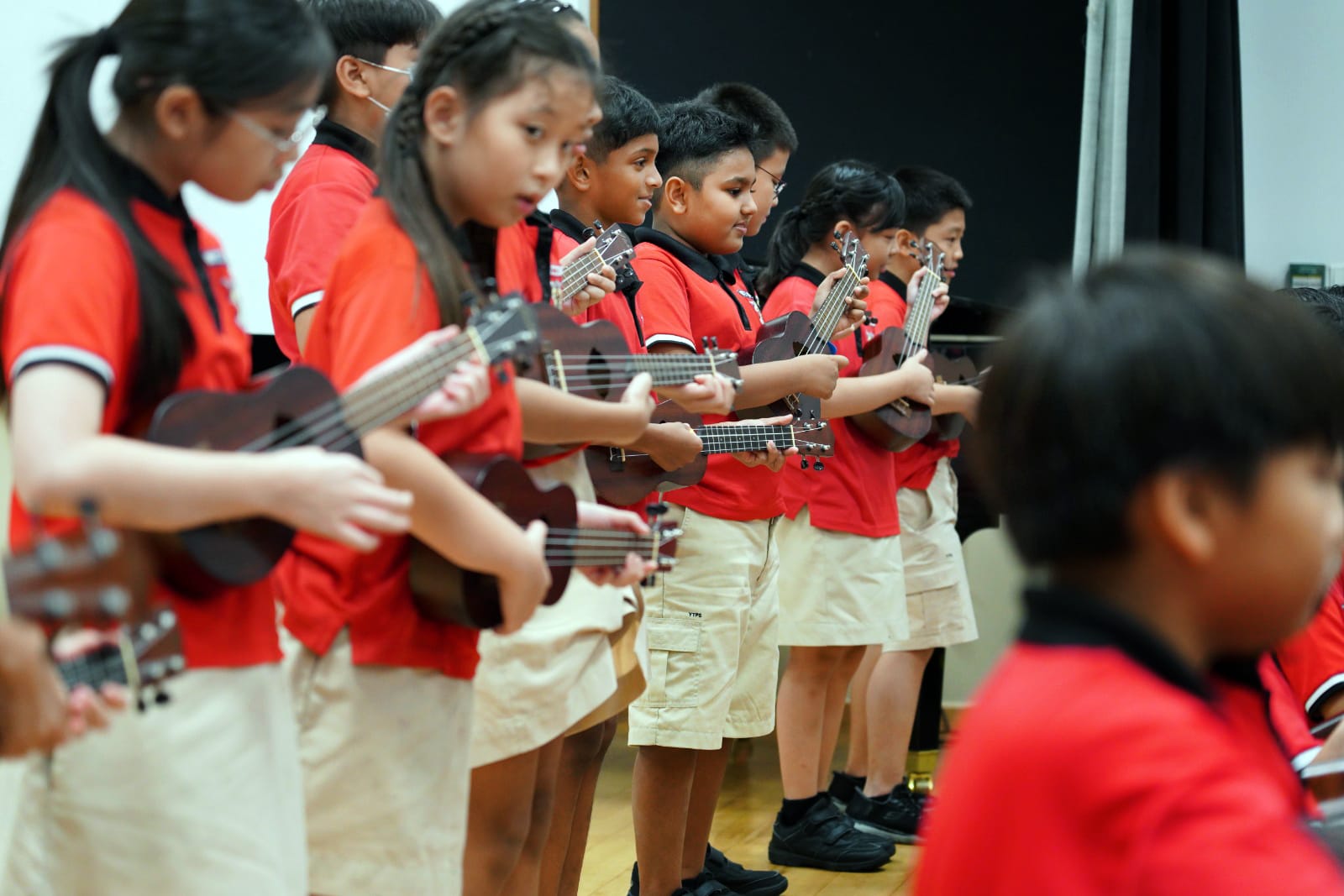 Row of schoolchildren in red shirts and khaki bottoms playing ukuleles on an indoor stage