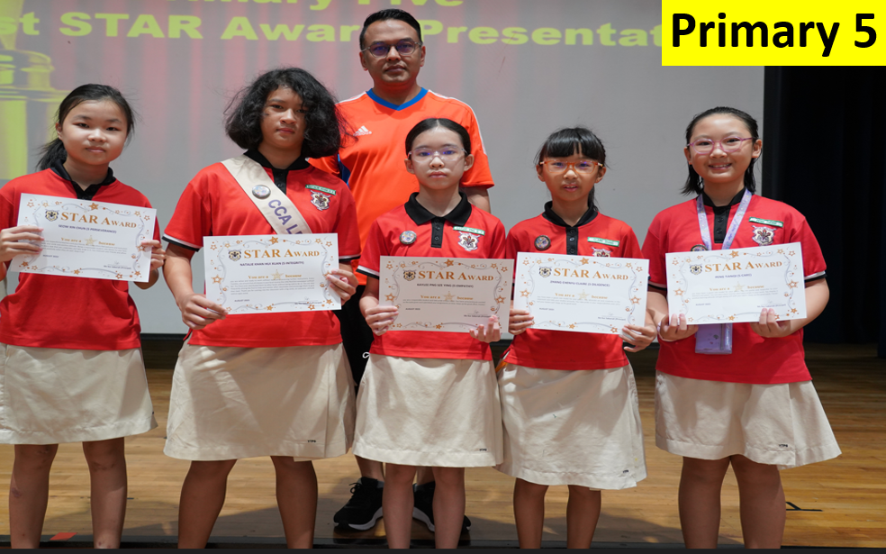 Five Primary 5 students in red uniforms hold Star Award certificates, standing with a male teacher on a stage.