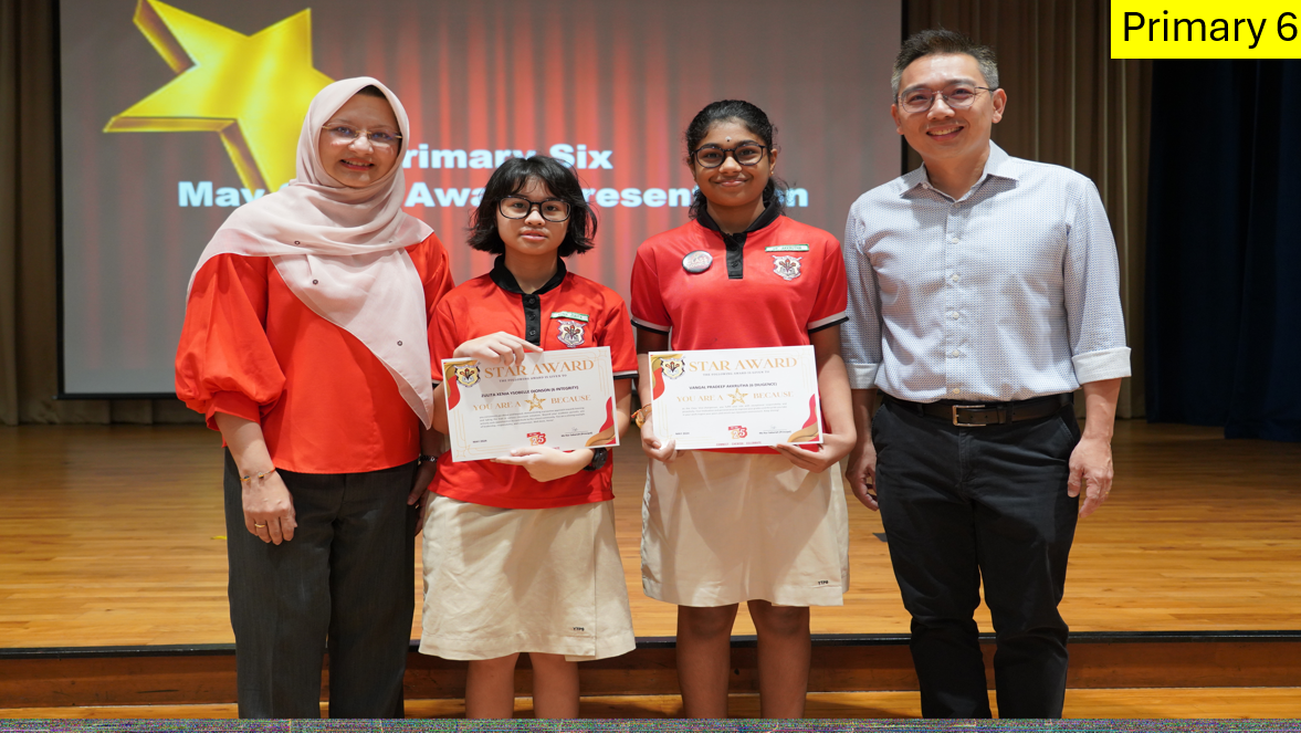 Two students in red uniforms hold Star Award certificates, flanked by two adults on a stage at a Primary Six award ceremony.