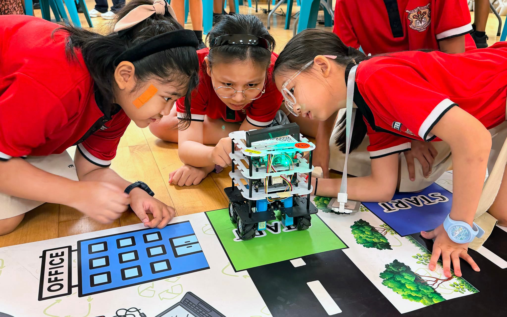 Three girls in red uniforms crouch over a small wheeled robot on a printed road mat.