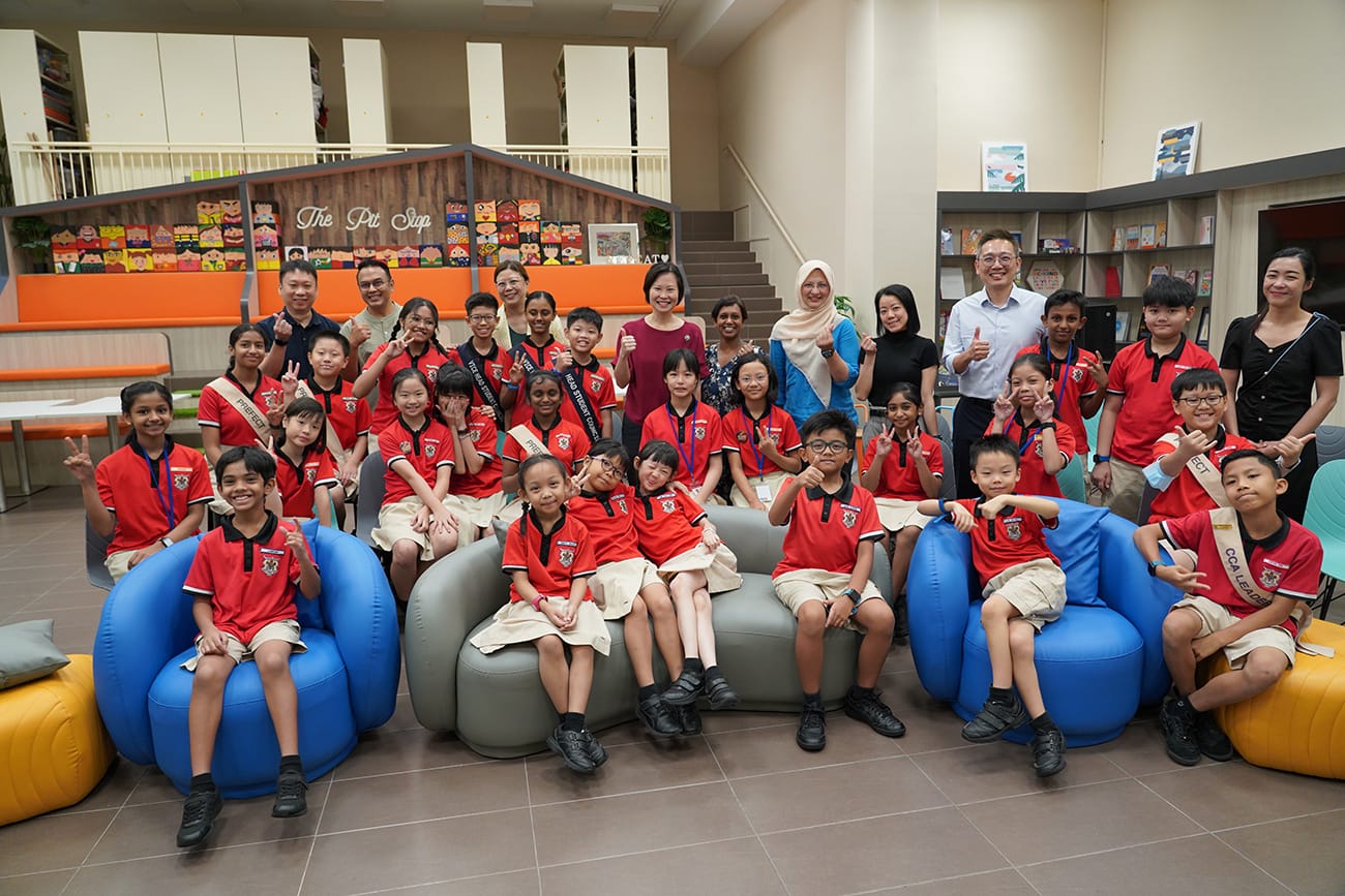 A large group of primary school children in red uniforms pose with adults in a colorful school common area.