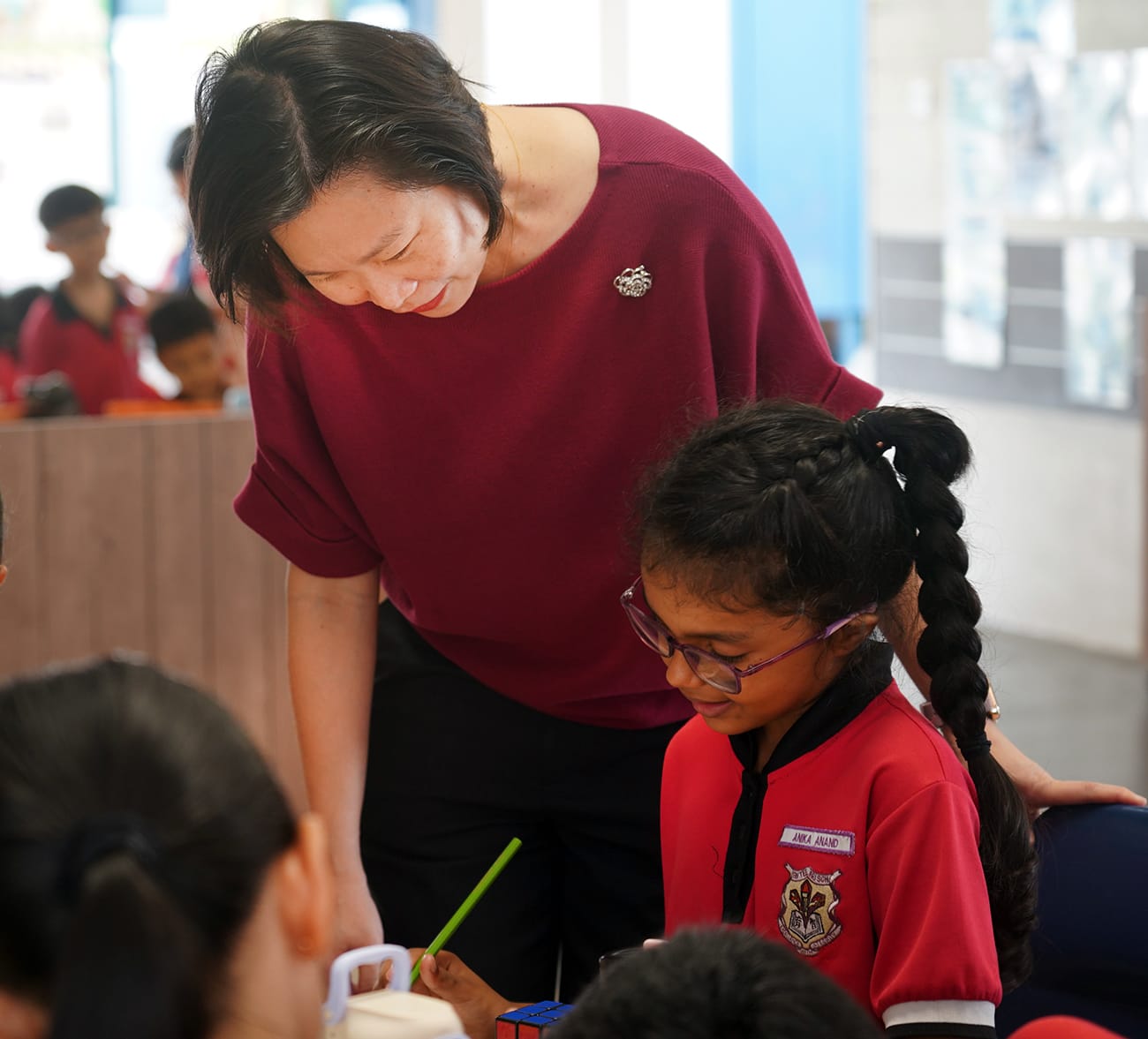 A woman in a red top leans over to assist a young girl in a red school uniform working with a Rubik's cube.