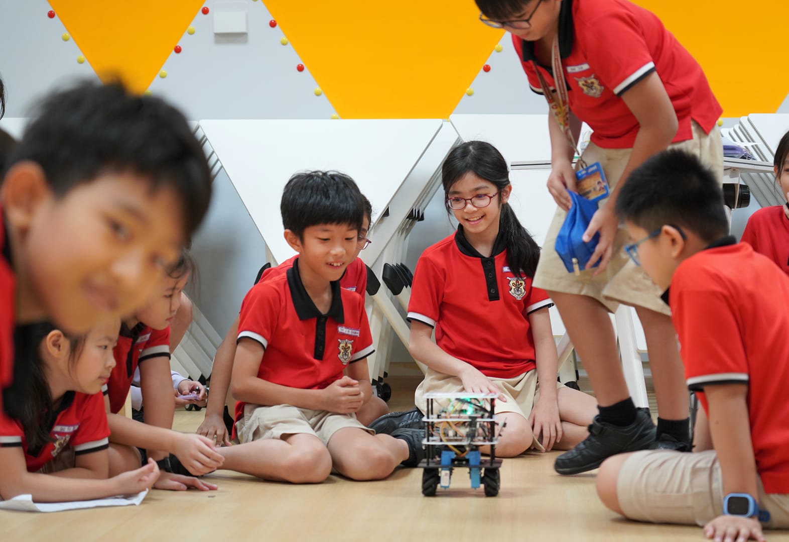 Several children in red school uniforms sit on a floor, gathered around a small wheeled robot they are examining.
