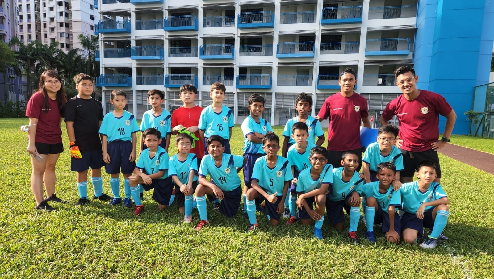 A youth football team in teal jerseys poses with two coaches on a grass field in front of a blue apartment block.