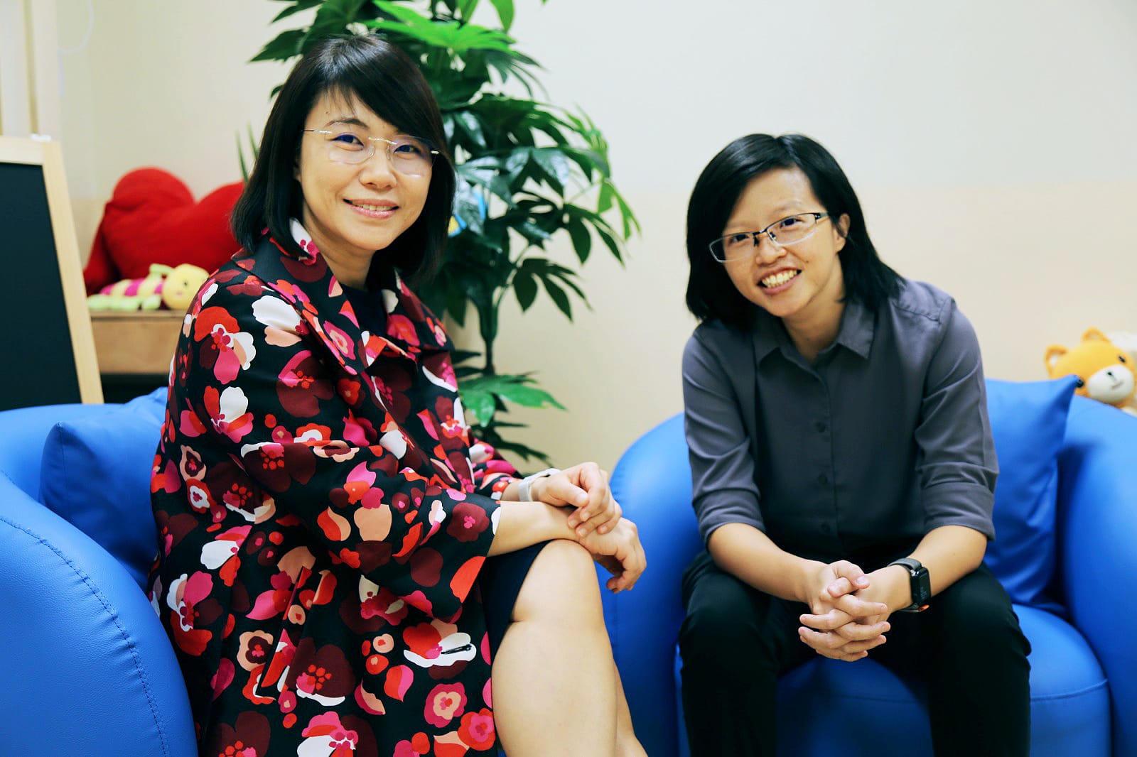 Two smiling women wearing glasses sit on blue chairs indoors, one in a floral dress, the other in a gray shirt.