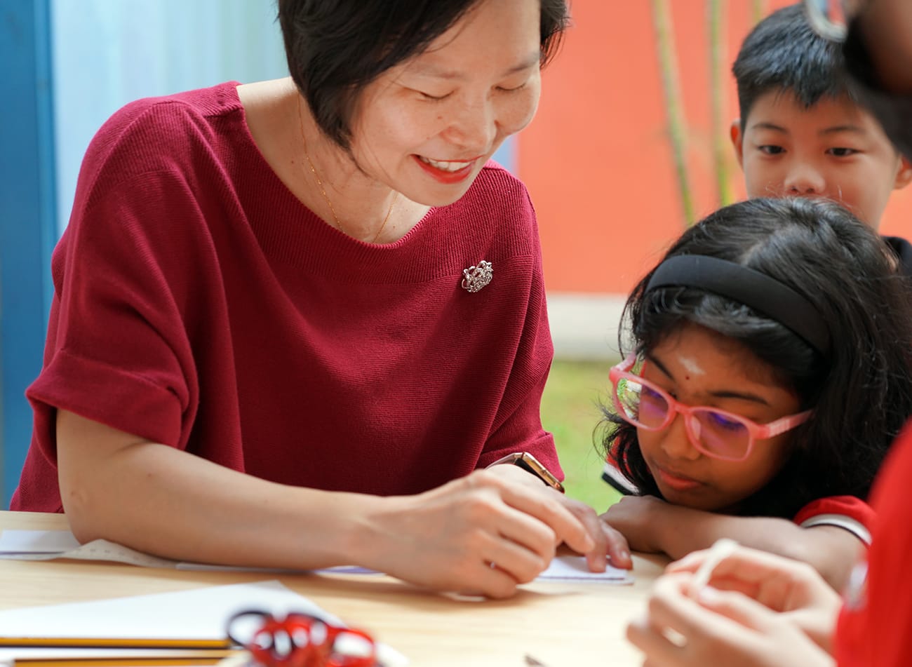 A smiling woman in a red top writes at a table while a girl with pink glasses and other children look on.