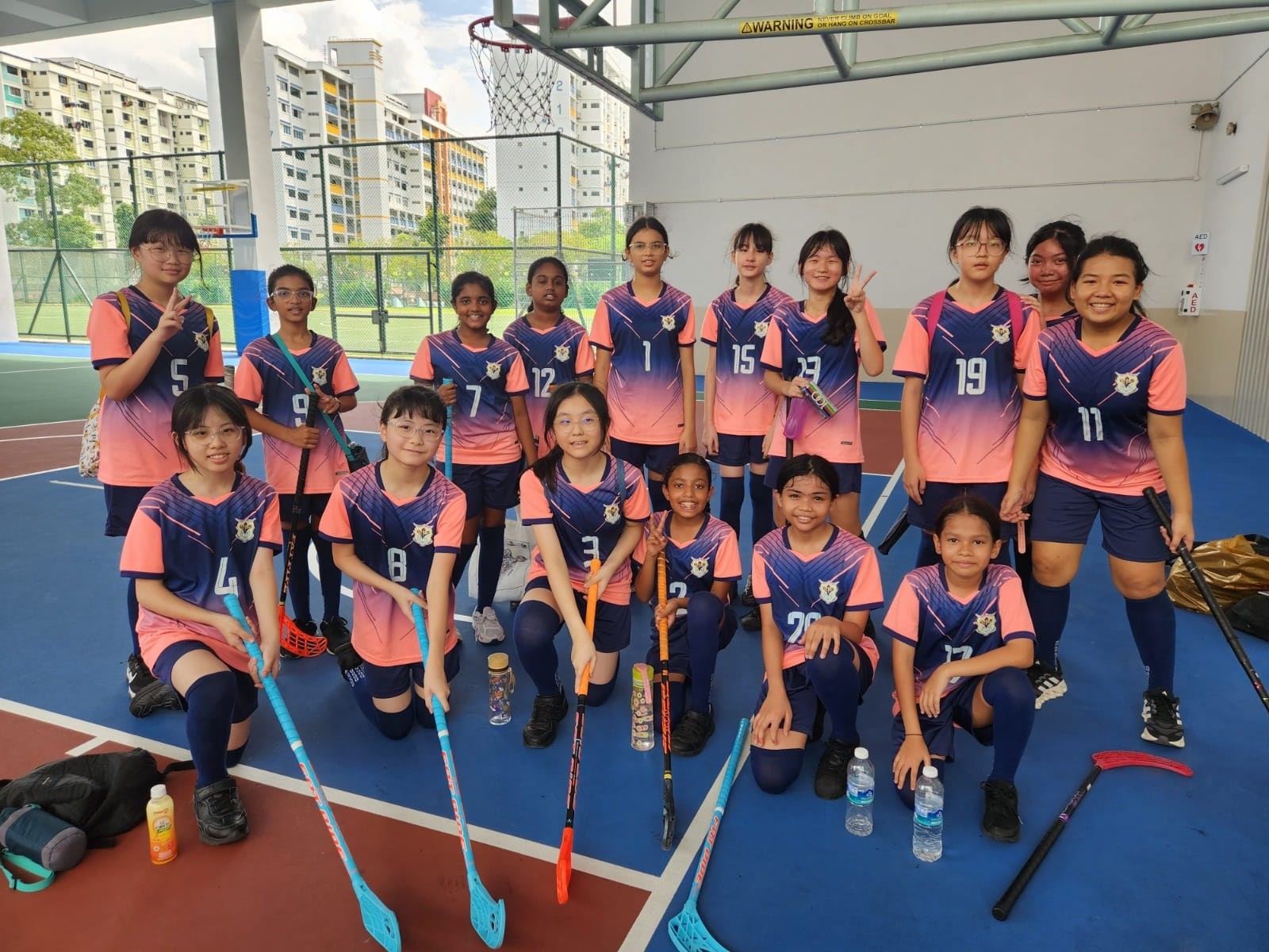 A girls floorball team in pink and navy uniforms posing with sticks on an outdoor covered court.