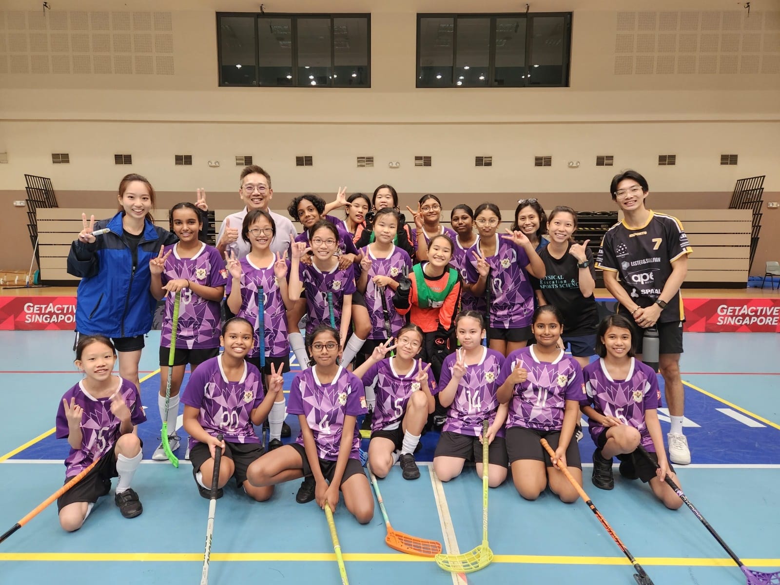 A girls floorball team in purple jerseys poses with coaches indoors, holding sticks and making peace signs.