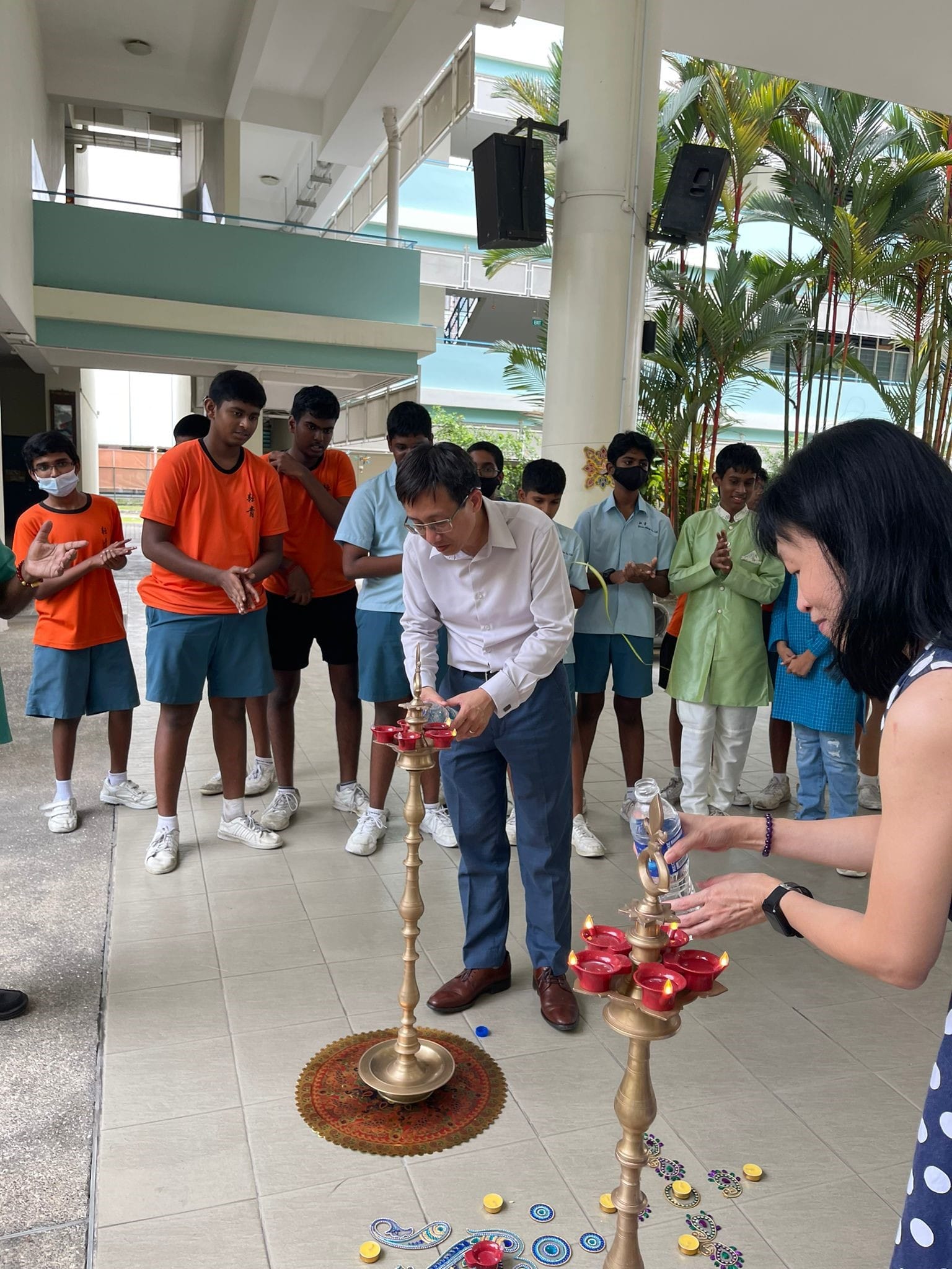 Two adults light brass oil lamps during a Diwali ceremony as a group of students watch and clap.