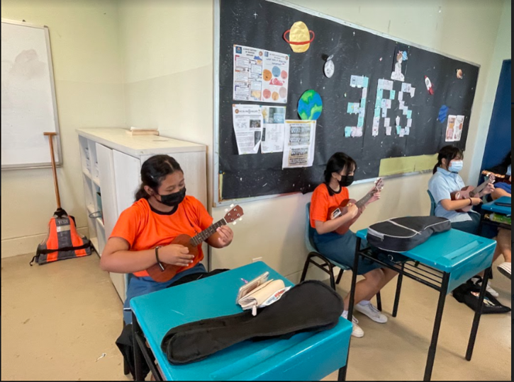 Students wearing masks sit at desks playing ukuleles in a classroom with a space-themed bulletin board.