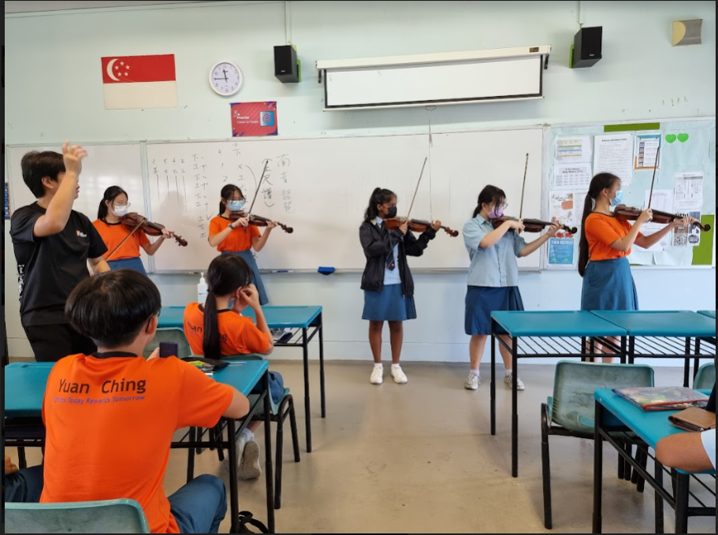 Students in a classroom stand in a row playing violins while a teacher conducts from the side and classmates watch from their desks.