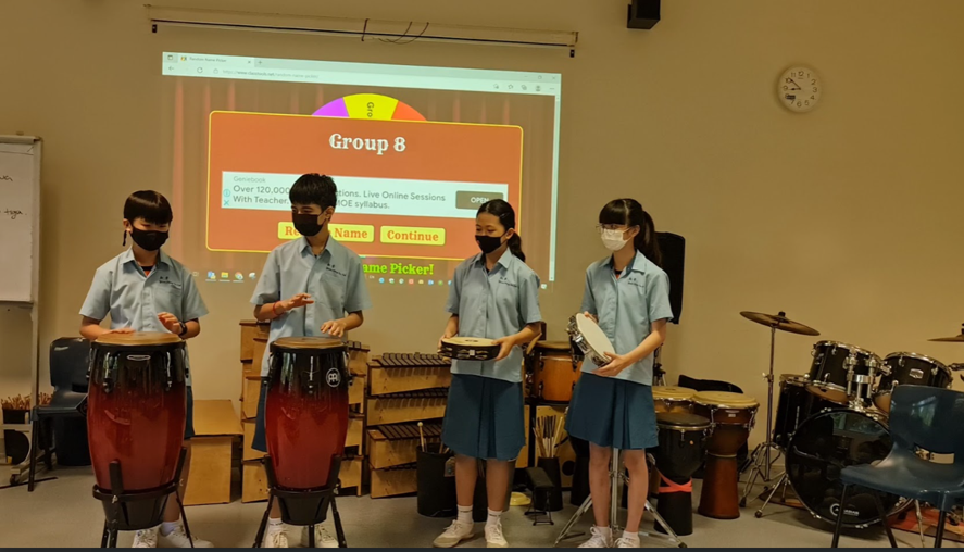 Students in school uniforms play percussion instruments like conga drums and tambourines during a music class presentation, with a screen displaying “Group 8” in the background.