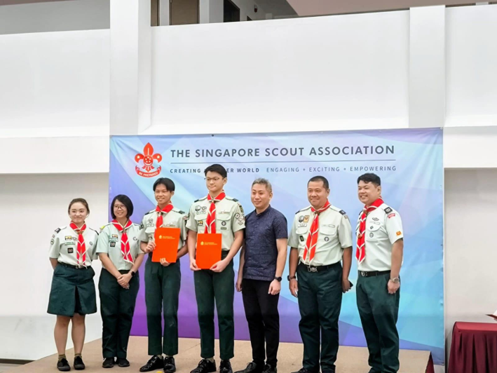 Scouts and adults pose on stage with certificates in front of a Singapore Scout Association banner during an award ceremony