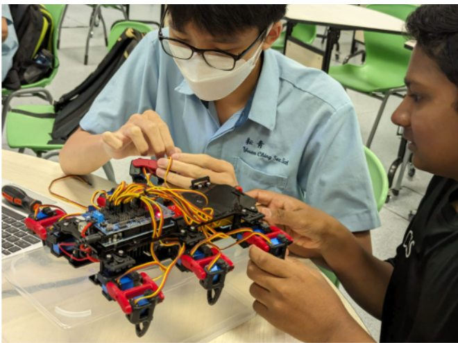Two students assemble a multi-legged robot with colorful wires and servo motors at a classroom table.