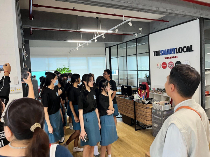 Photograph of a group of students wearing black shirts and blue skirts standing in an office space with a sign reading "THESMARTLOCAL SINGAPORE" on a glass partition.