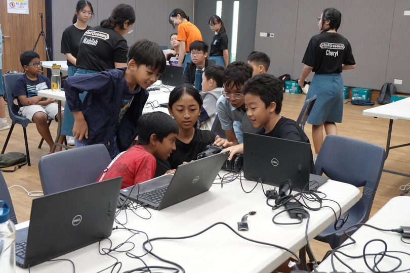Photograph of a group of children and teenagers gathered around laptops in a classroom or workshop setting, engaged in a collaborative activity.