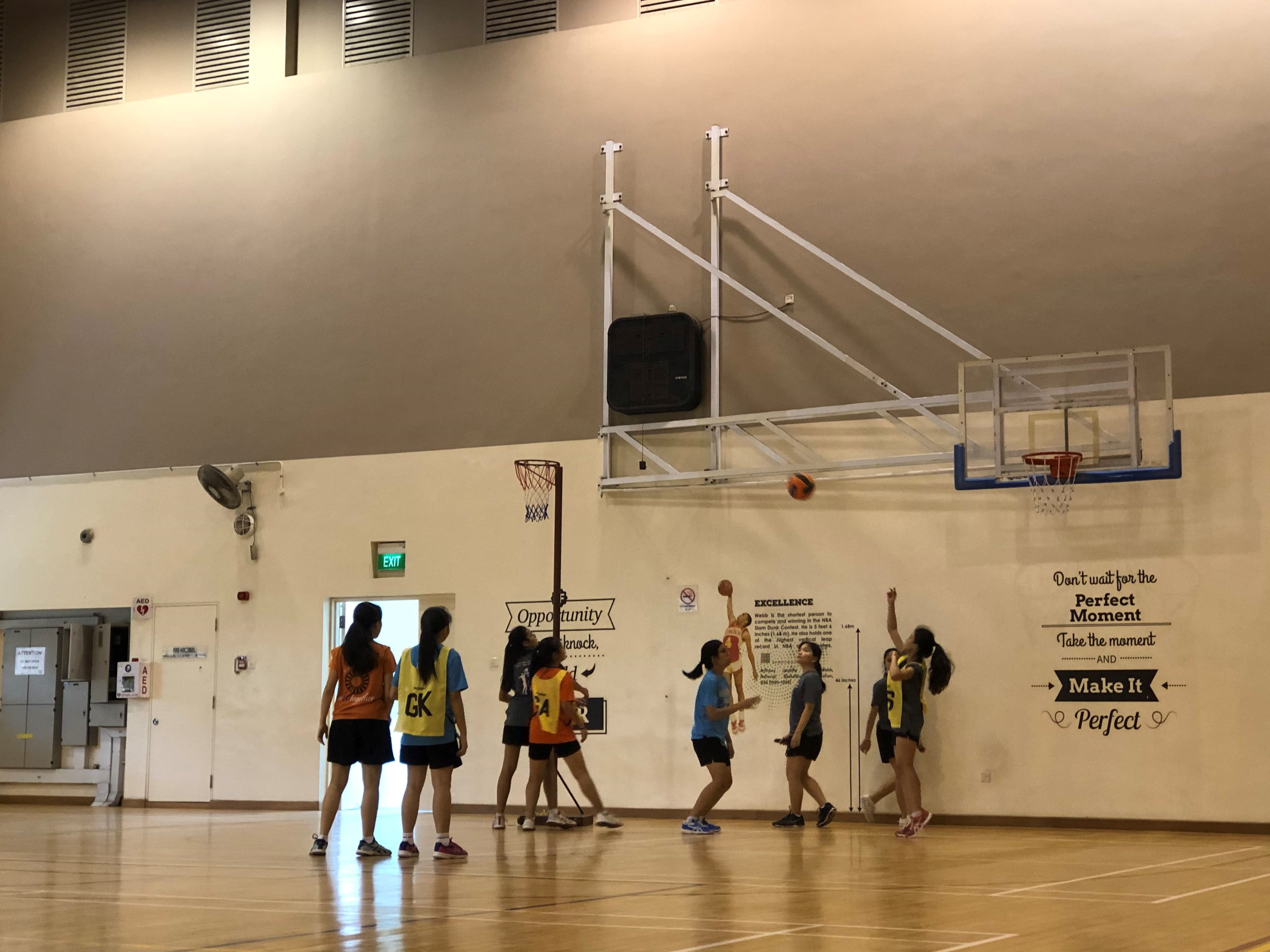 Photograph of a girls' basketball game in an indoor gym with six players near the hoop, one player in blue shooting the ball towards the basket. Wall behind hoop features motivational quotes in black text, including "Don't wait for the perfect moment, take the moment and make it perfect."