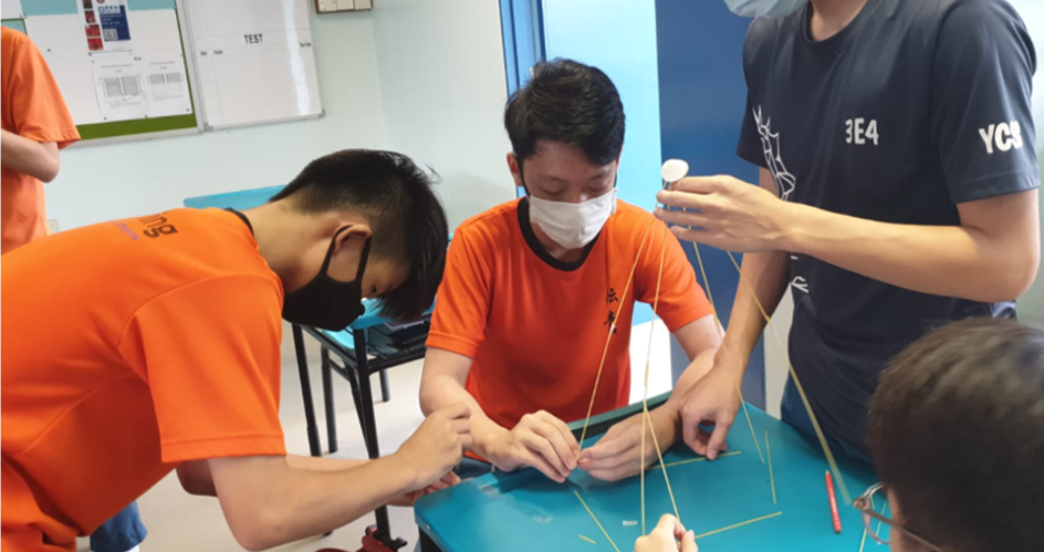 Students wearing masks and orange shirts work together building a structure with thin sticks on a blue table.