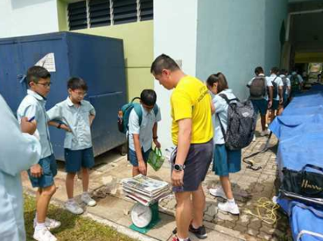 Students weighing a stack of newspapers on a scale during an outdoor environmental education activity at school.