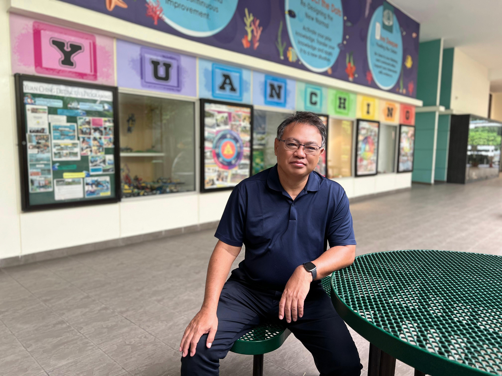 Man in navy polo shirt and glasses sits on a stool by a green table in front of Yuan Ching school display boards.