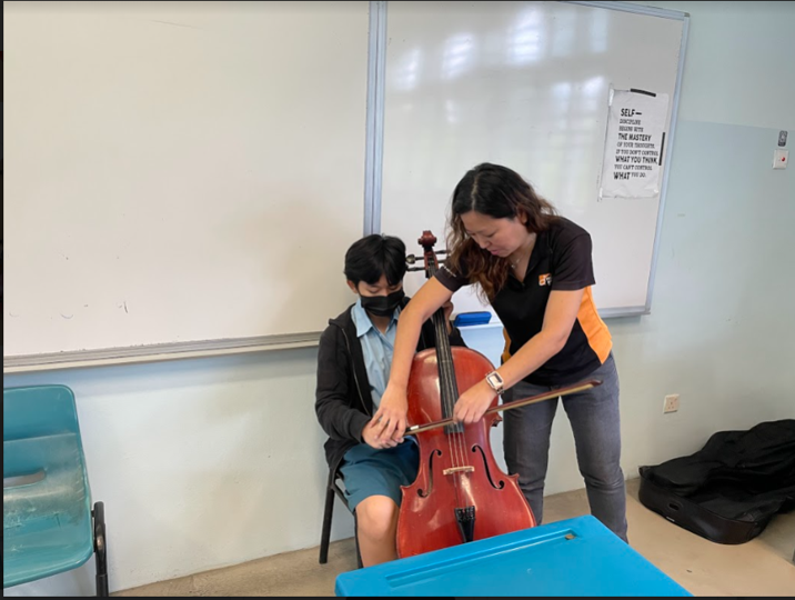 Teacher guides a seated student in proper cello playing technique in a classroom, demonstrating bow and hand positioning.