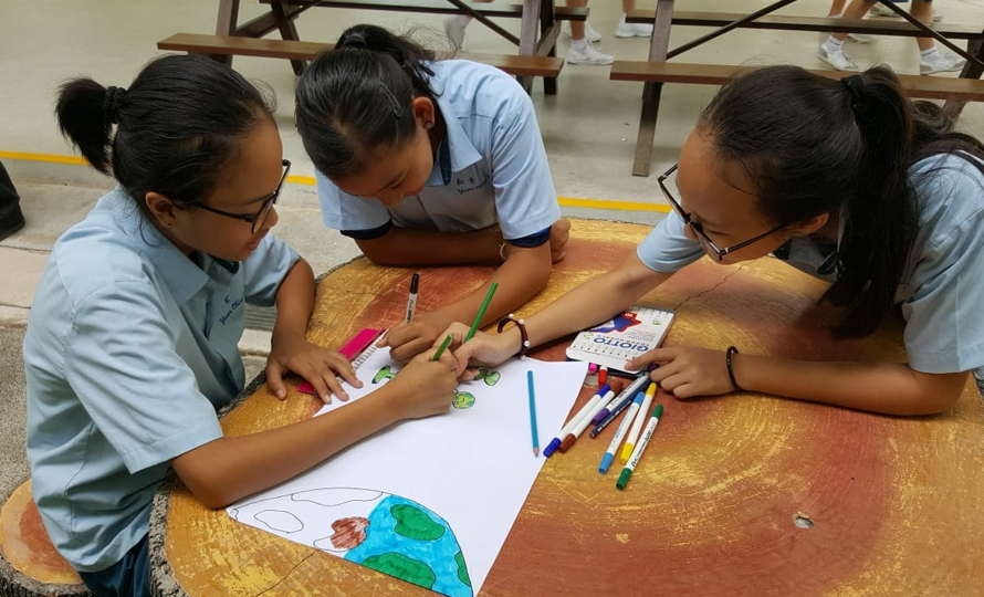 Students working together on an environmental-themed poster featuring the Earth, using coloured markers at an outdoor table.