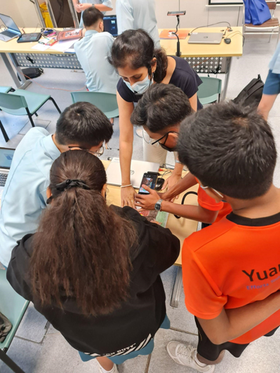 Photograph of a group of students gathered around a table in a classroom, collaborating on a project involving a smartphone and electronic components.