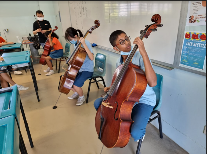 Students sit in a classroom practicing cello, holding their instruments upright while a teacher supervises and assists in the background.