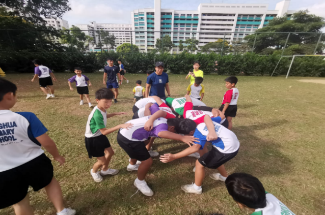Children in school uniforms playing a rugby scrum on a grass field with tall buildings in the background.