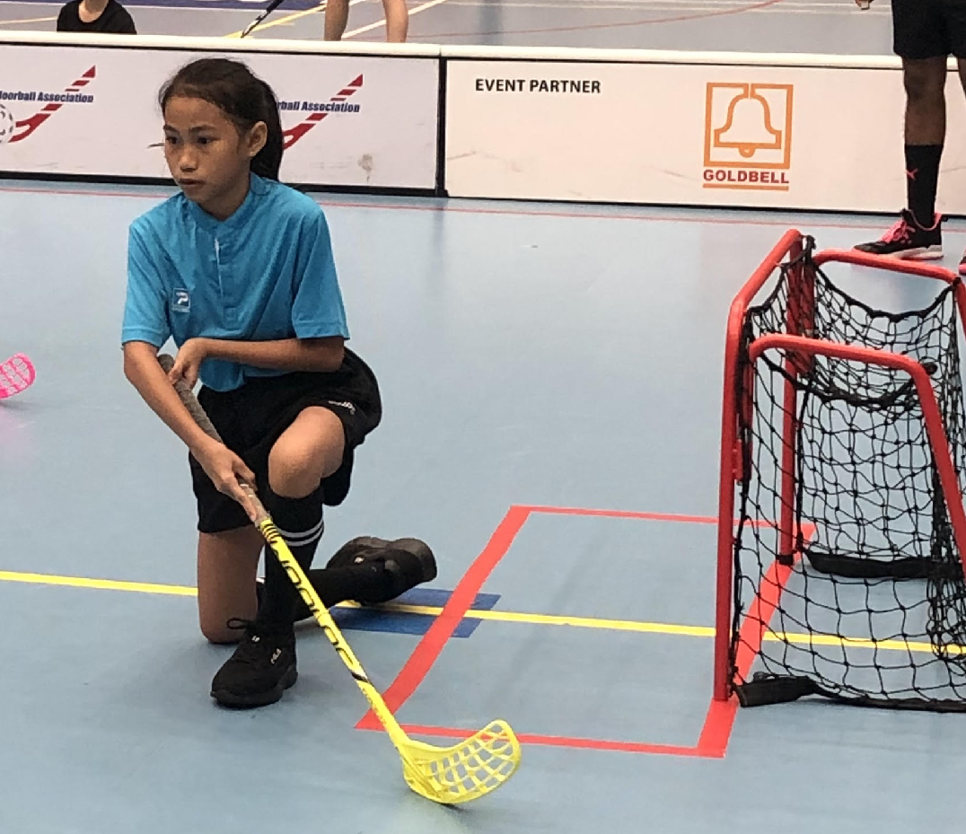 A young girl in a blue jersey kneels on an indoor court holding a yellow floorball stick near a small red goal.