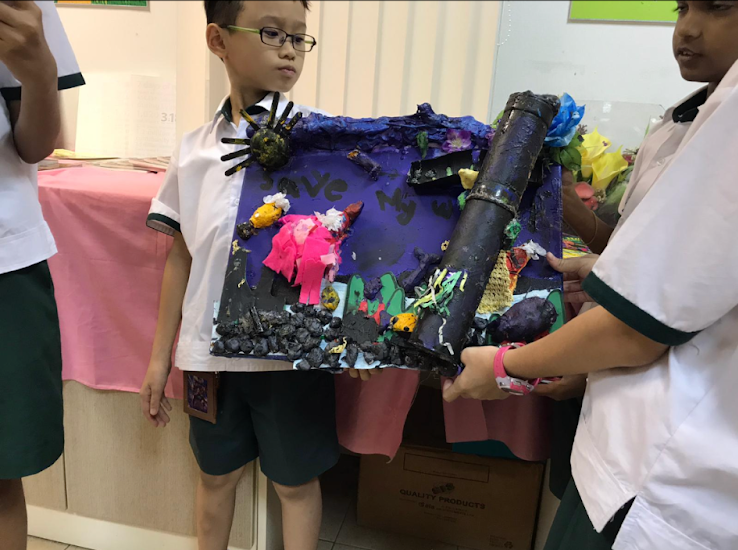 Two schoolboys in white uniforms hold a colorful diorama with a large black pipe and debris.