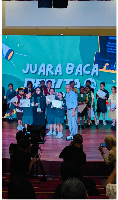 Students holding certificates on stage at a Juara Baca Berita competition, with an adult presenter and teal backdrop.