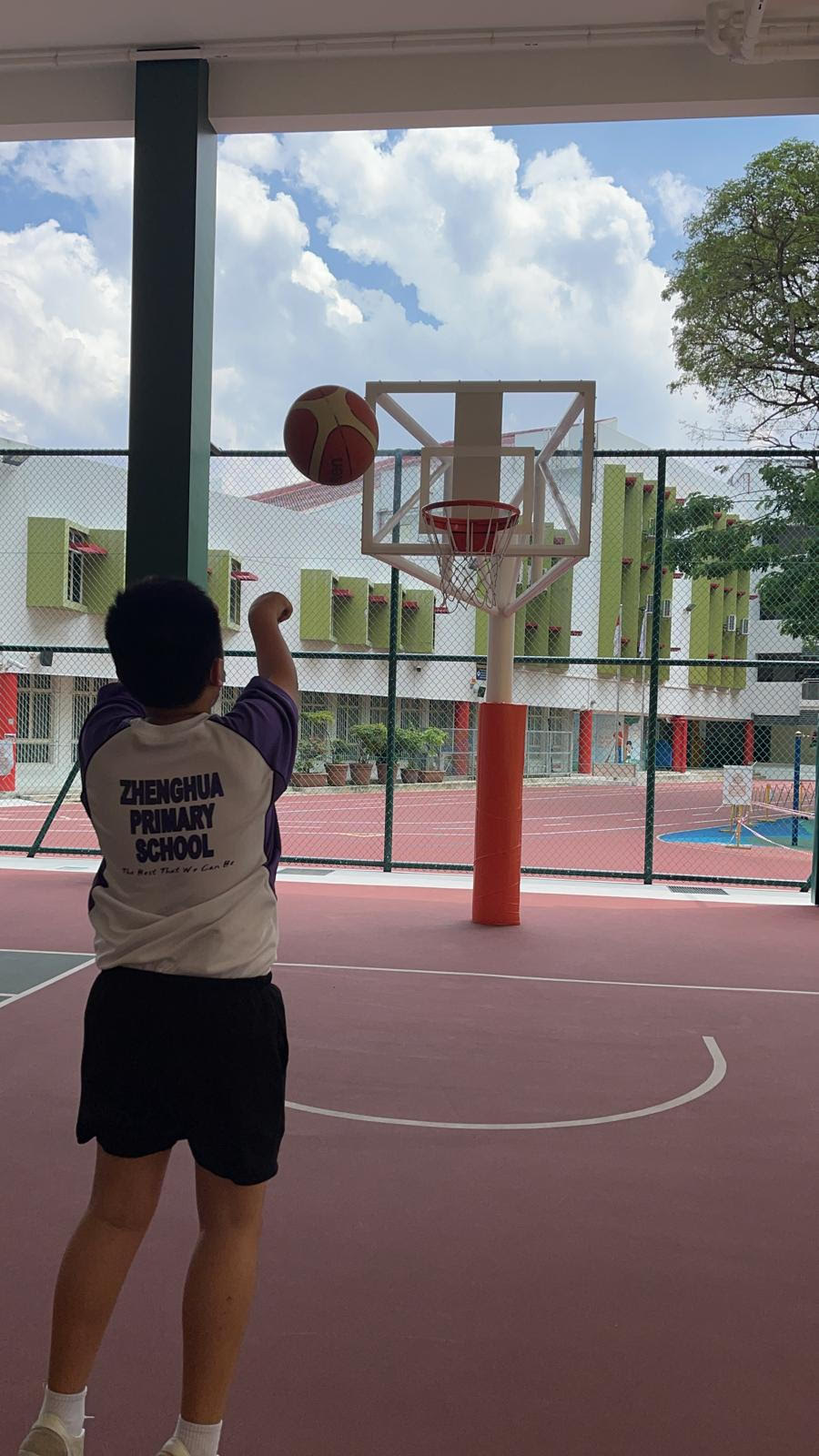A child in a Zhenghua Primary School shirt shoots a basketball toward a hoop on an outdoor court.
