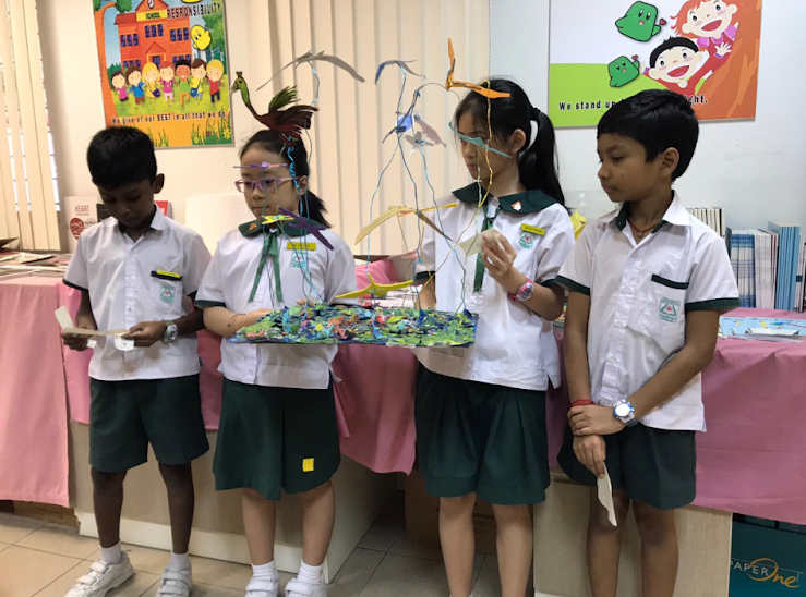 Four schoolchildren in uniforms stand by a table, one holding a colorful model with tall wire shapes.