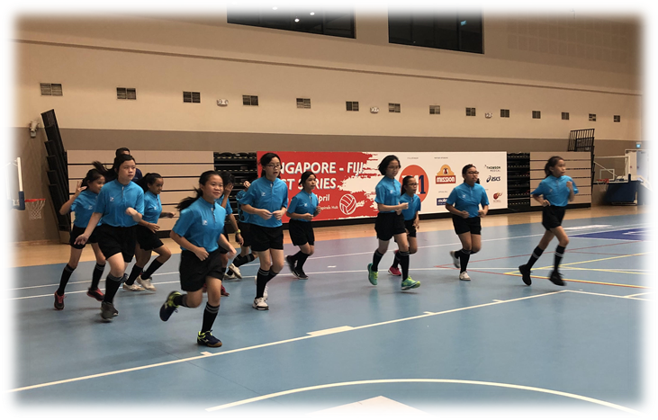 Girls in blue jerseys and black shorts run across an indoor court with a Singapore-Fiji series banner behind them.