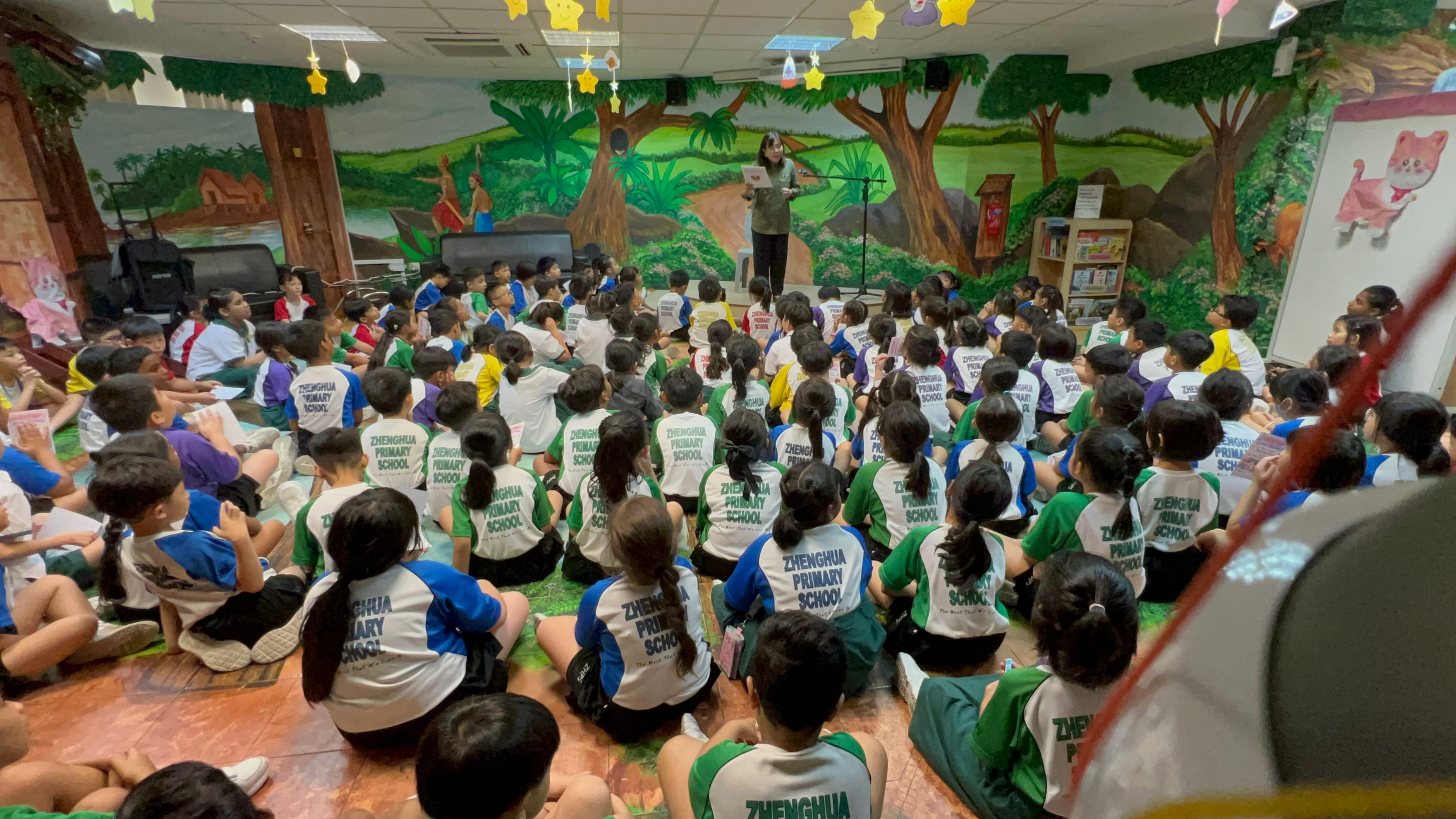 A teacher stands before many Zhenghua Primary School students seated on a floor in a decorated library room.