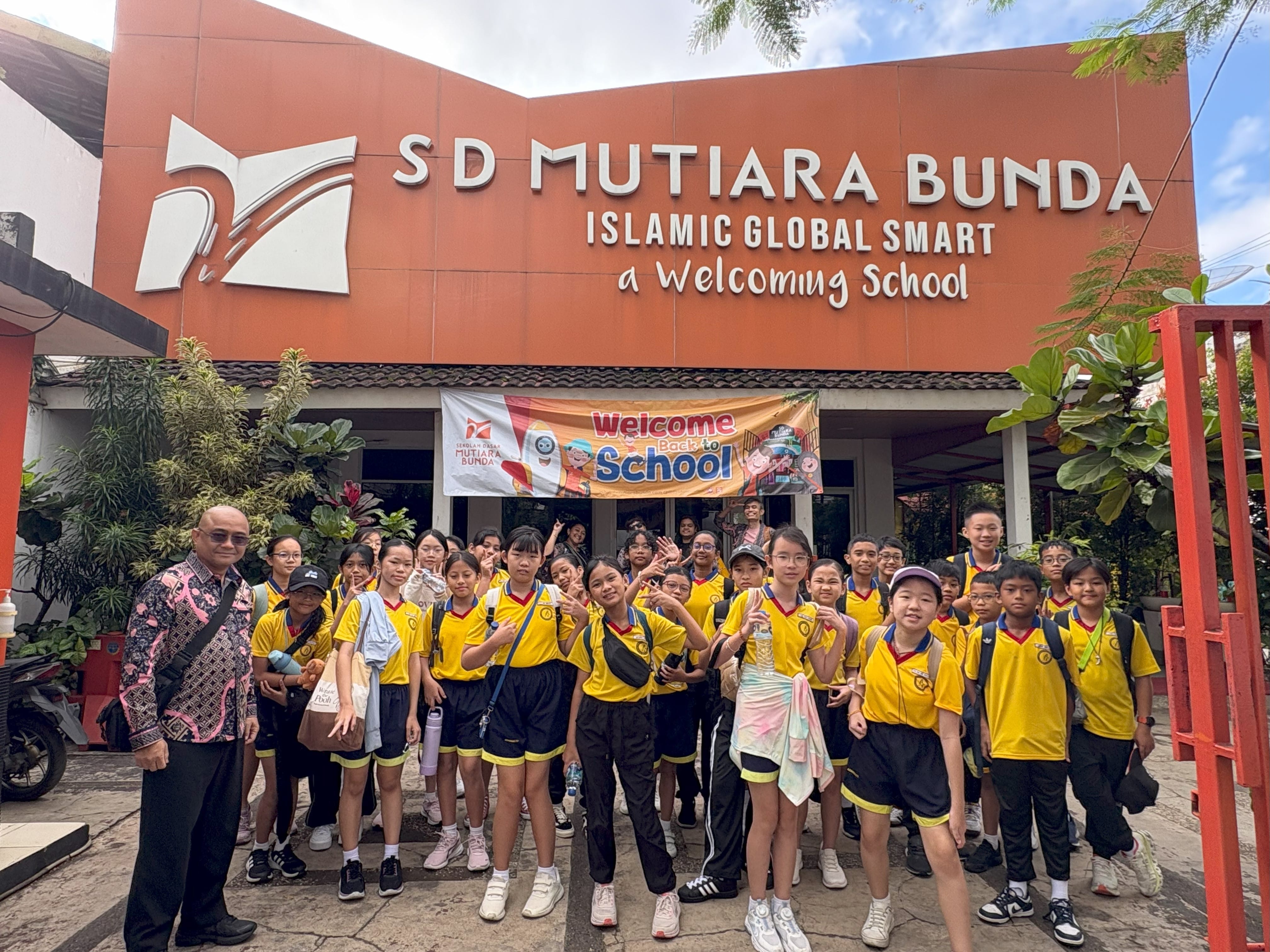 Students in yellow uniforms pose with an adult outside SD Mutiara Bunda school under a Welcome Back to School banner.