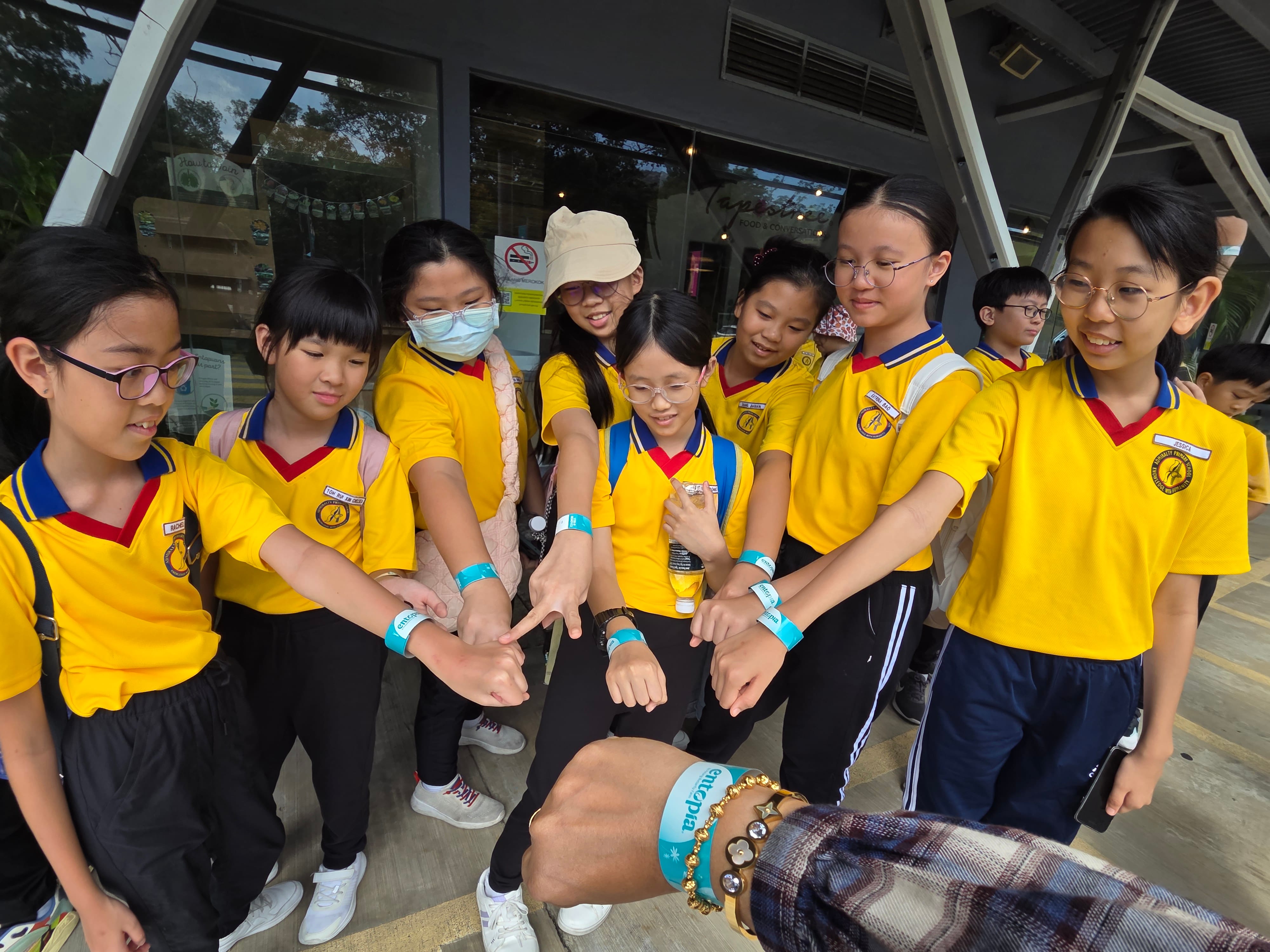 A group of children in yellow school uniforms pointing at teal wristbands on their wrists outdoors.