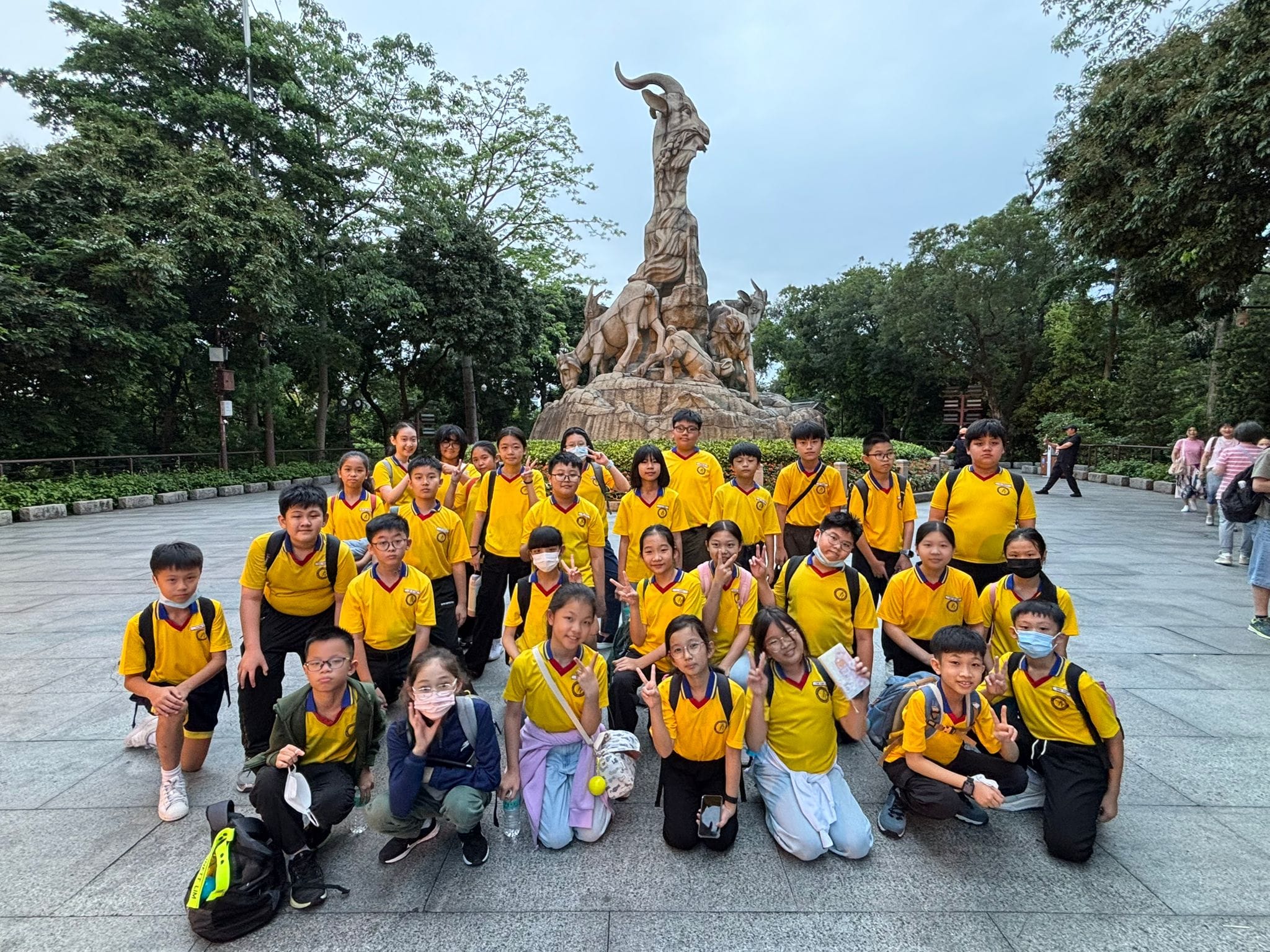 A large group of children in yellow uniforms pose in front of the Five Rams stone sculpture in a park.