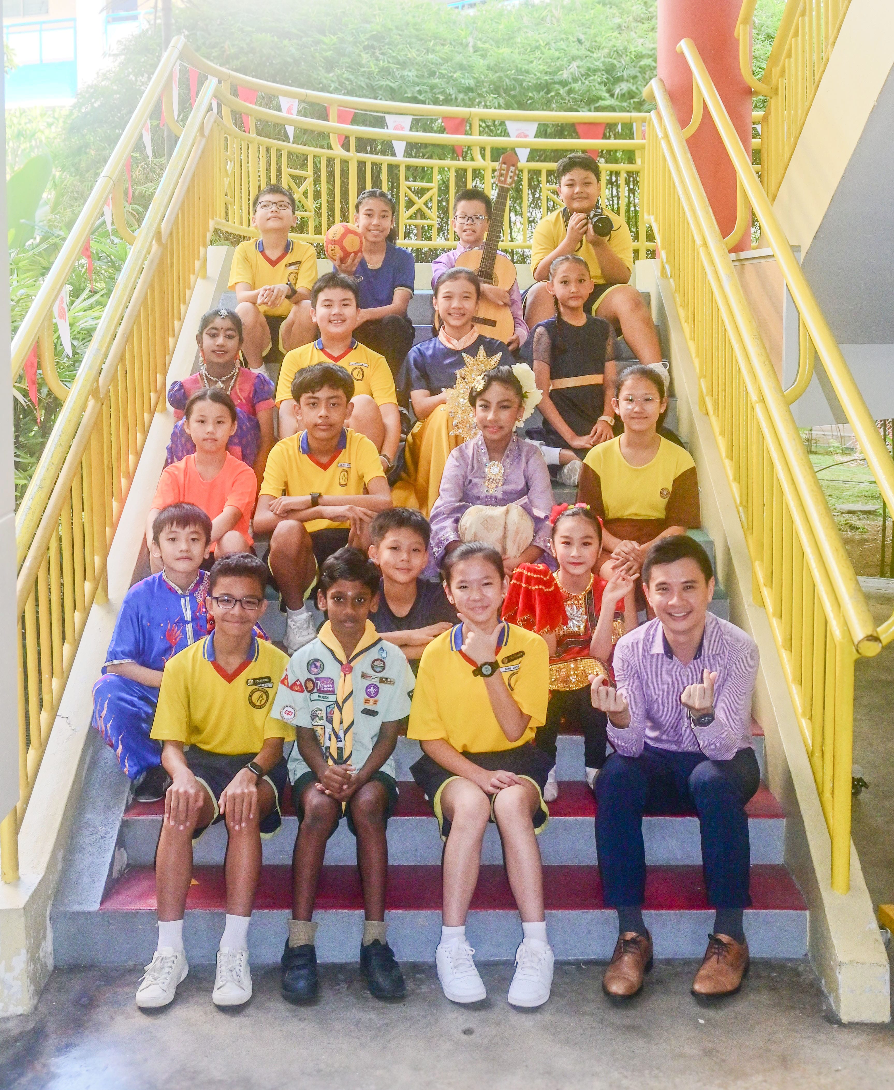 A group of school children and an adult teacher pose on outdoor stairs with yellow railings, wearing various uniforms and costumes.