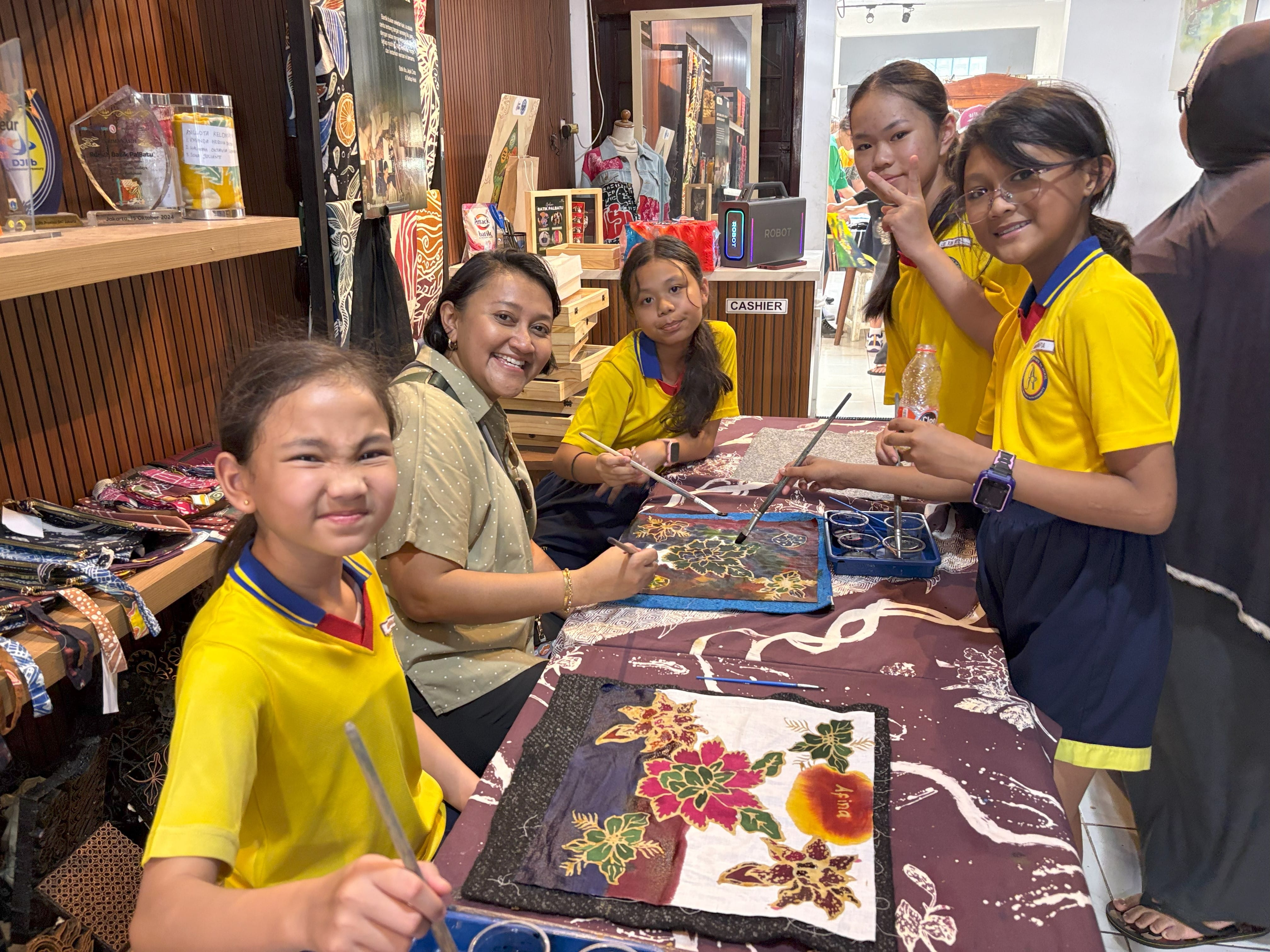 Four girls in yellow uniforms and an adult woman sit at a table doing batik fabric painting in a craft shop.
