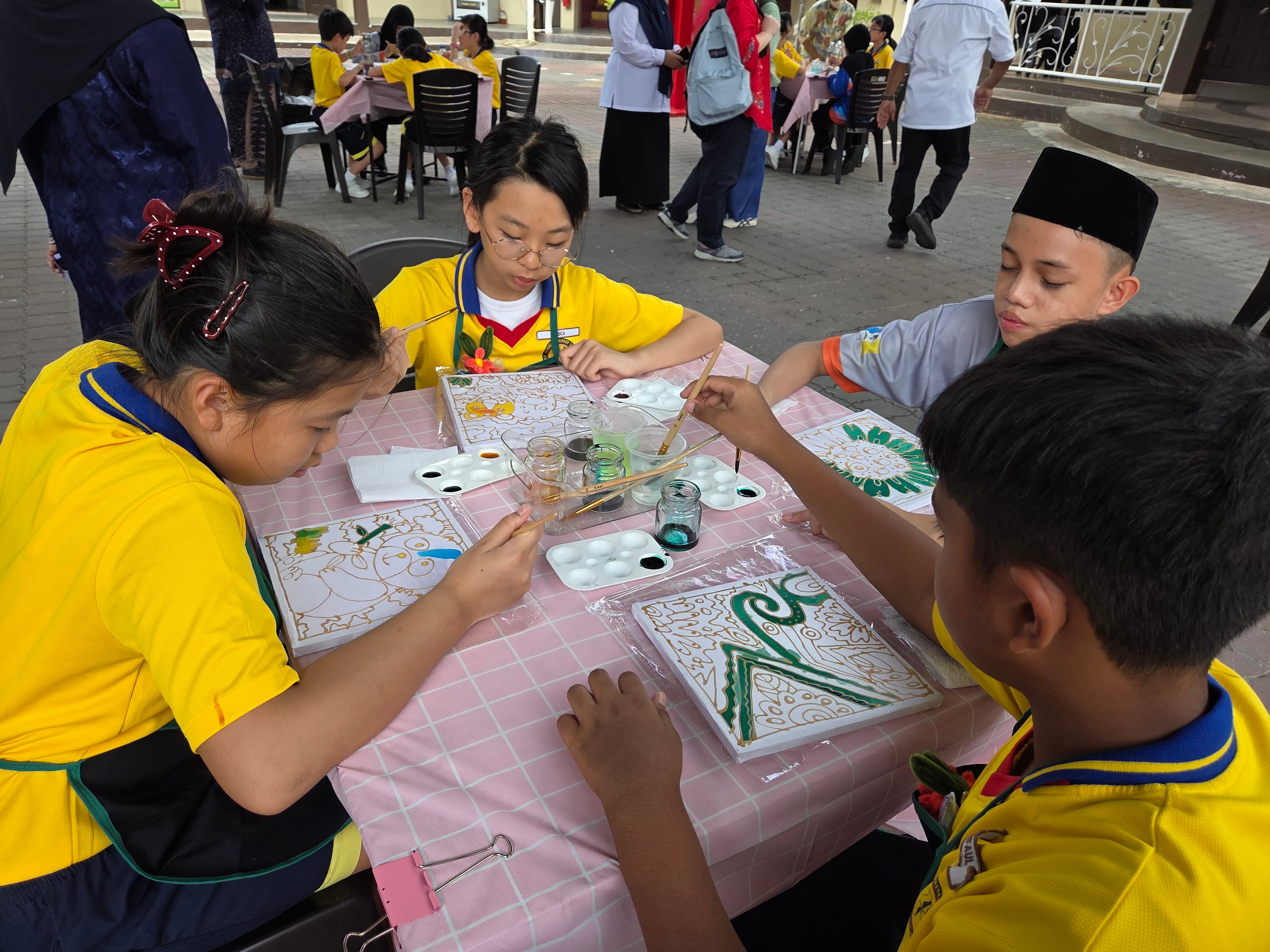 Four children in yellow uniforms paint decorative art panels at an outdoor table with paint jars and palettes.