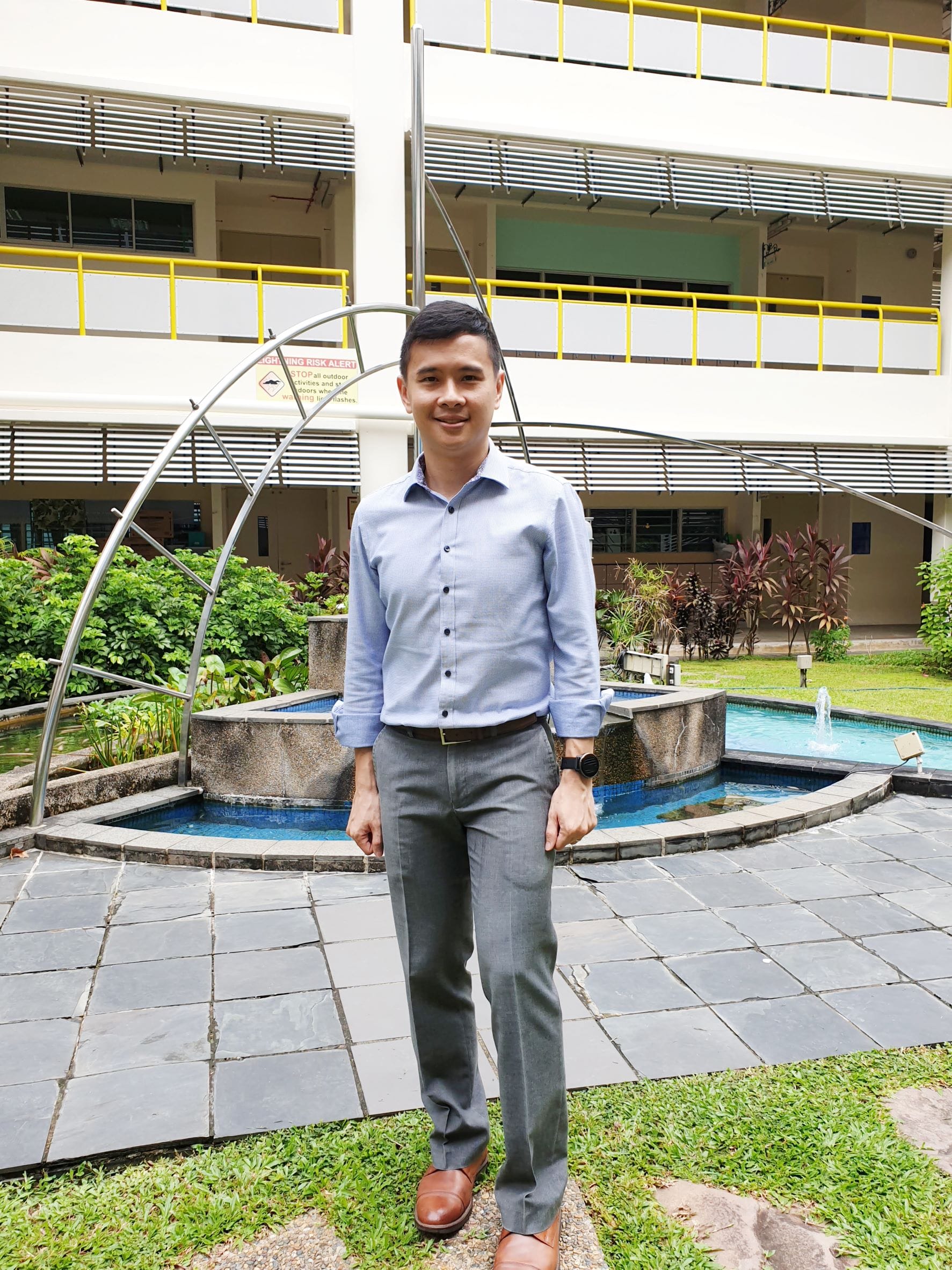 A young man in a blue shirt and grey trousers stands smiling in front of a fountain courtyard beside a multi-storey building.