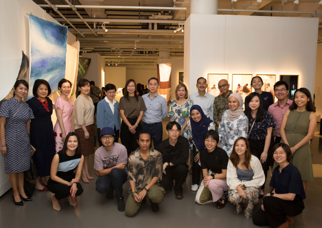 A large group of people posing together inside an art gallery with hanging fabric artworks and framed pictures on walls.
