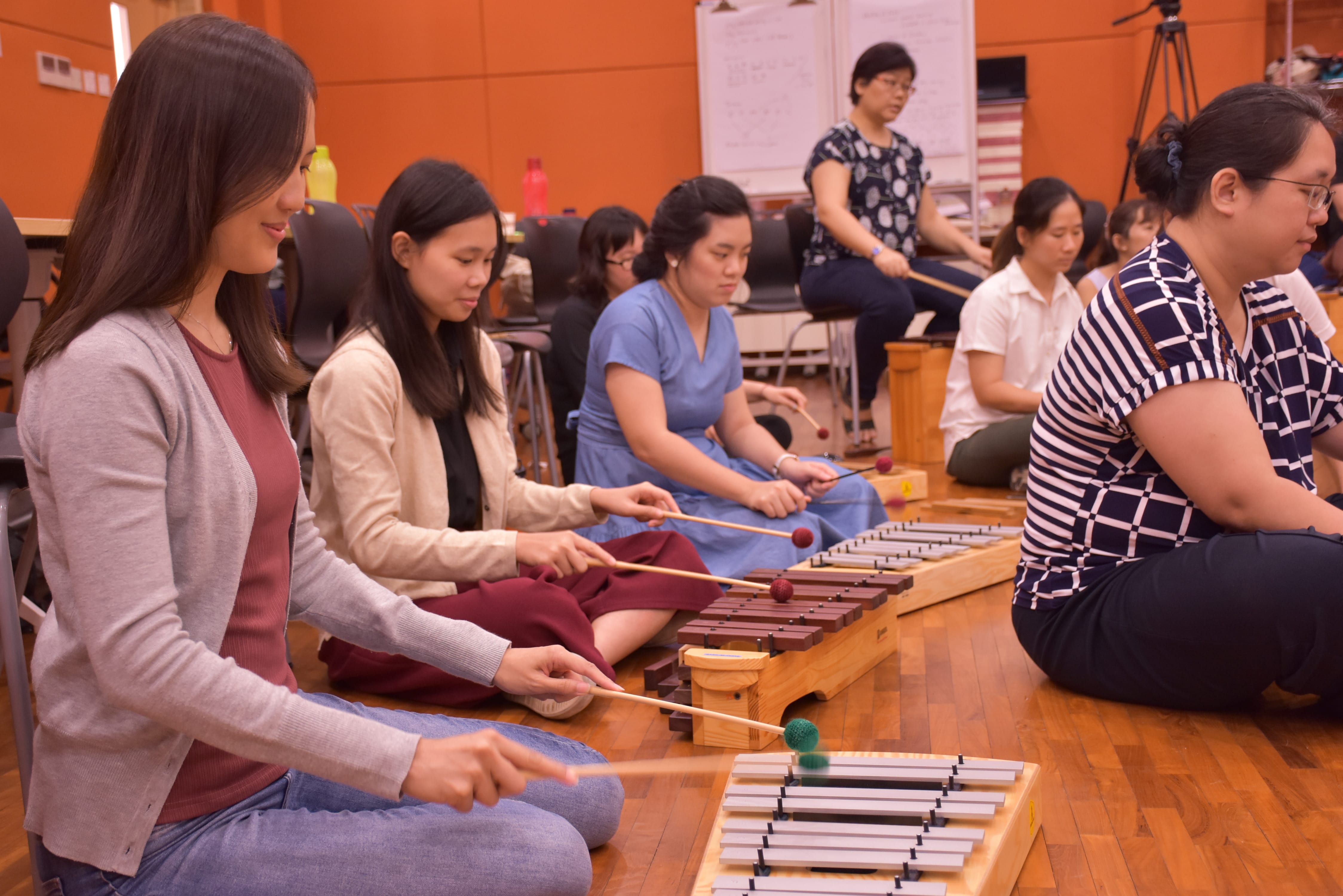 A photo taken during the Orff workshop. Women seated on floor playing xylophones with mallets in a group music activity.