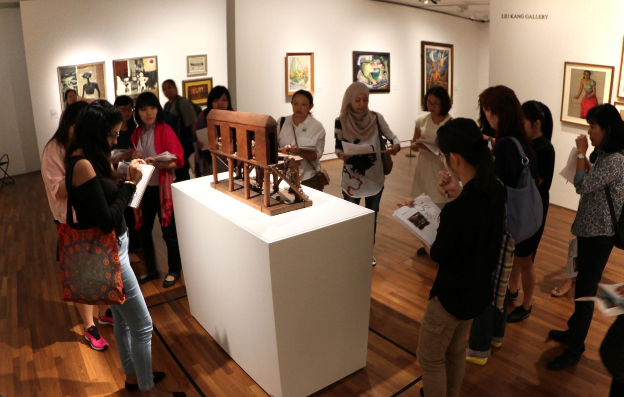 A group of art teachers gather around a wooden sculpture on a white pedestal in the Liu Kang Gallery.
