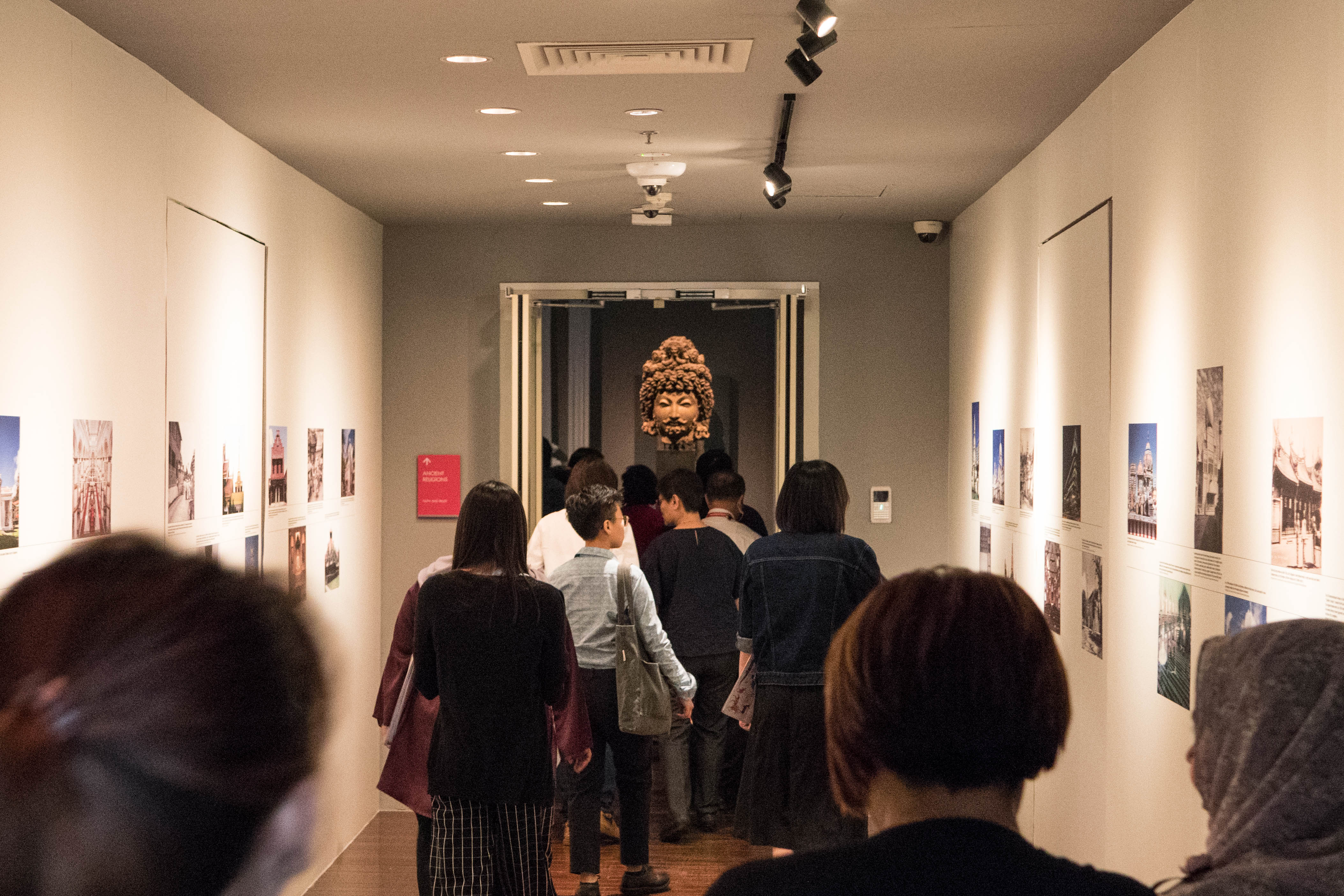 A group of visitors walk through a museum corridor with photos on walls, toward a sculpted head displayed in a doorway.