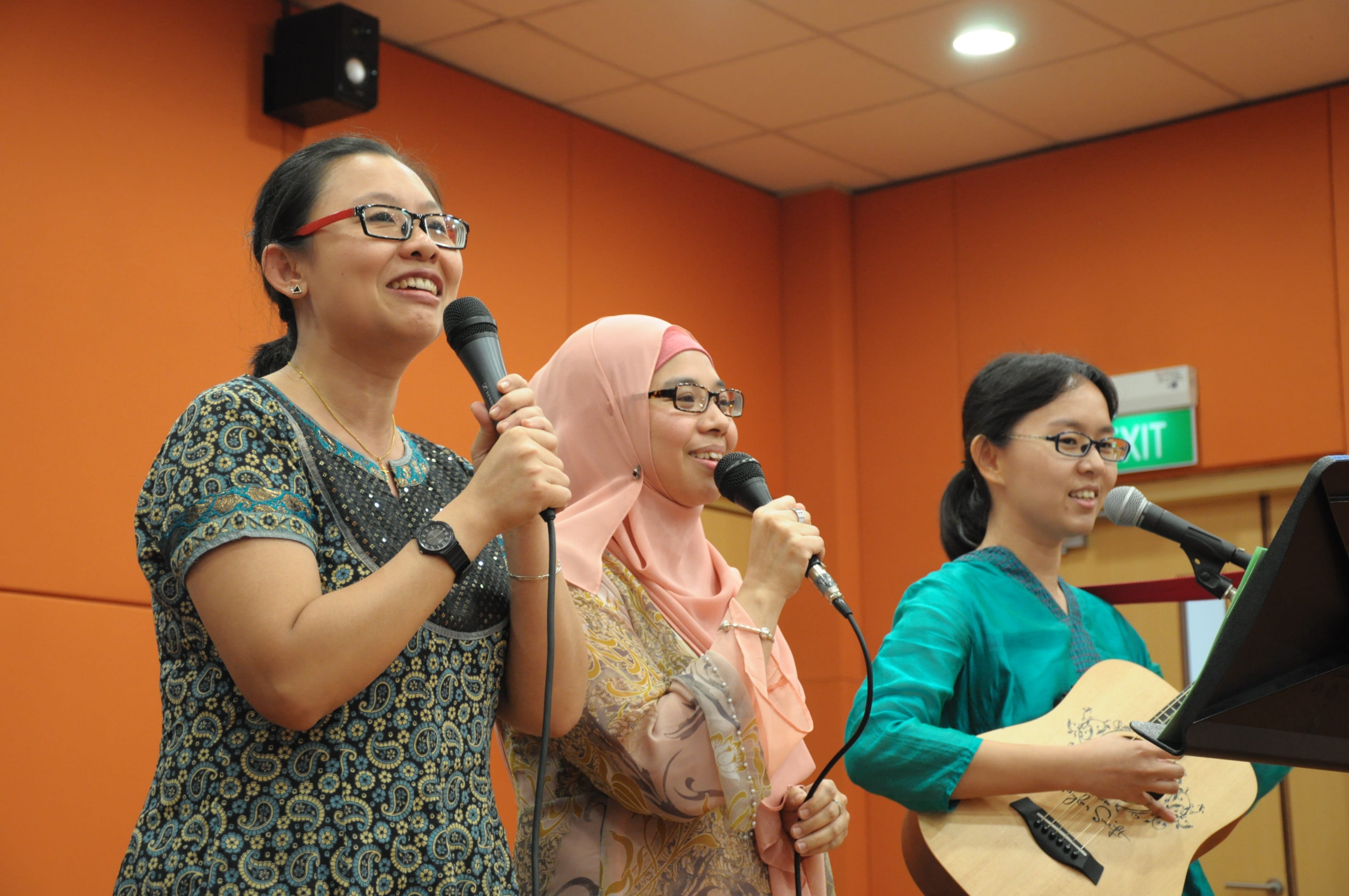 Photo taken during Music Teacher Practitioner Programme. Three women performing, two holding microphones and singing, one playing acoustic guitar, in an orange-walled room.